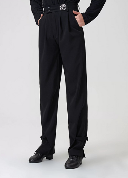 A model wearing black BOBO DANCE ballroom dance pants with a matching black belt featuring a silver 'BD' logo, paired with a black long-sleeve top and black dance shoes, against a light gray studio background.