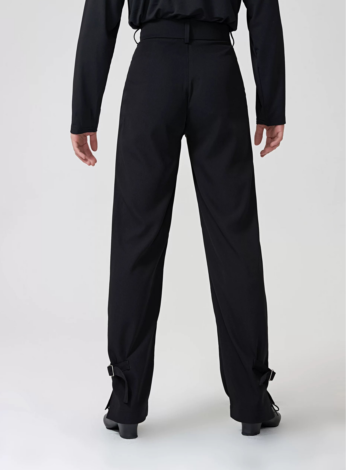 A model from the back wearing black BOBO DANCE dancewear, featuring a long-sleeve top and high-waisted, wide-leg pants with ankle straps, set against a plain light gray studio background.